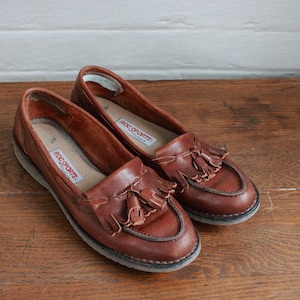 May include: A pair of brown leather loafers with tassels. The loafers have a white sole and the brand name "Roc Sports" is printed on the insole.