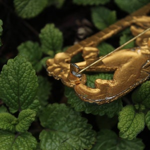 May include: A gold-toned metal brooch with a detailed design of a stylized animal. The brooch is lying on a bed of green leaves.