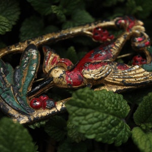 May include: A gold, red, and green metal brooch in the shape of a bird with leaves and berries. The brooch is on a bed of green leaves.