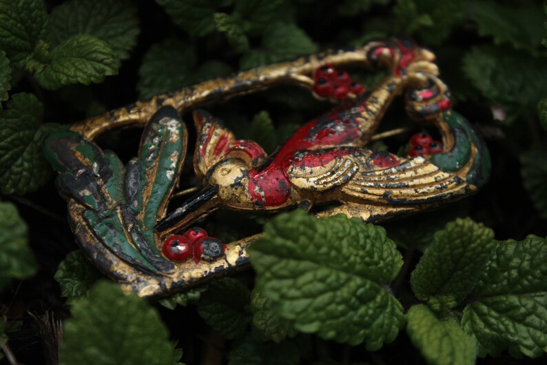 May include: A gold, red, and green metal brooch in the shape of a bird with leaves and berries. The brooch is on a bed of green leaves.