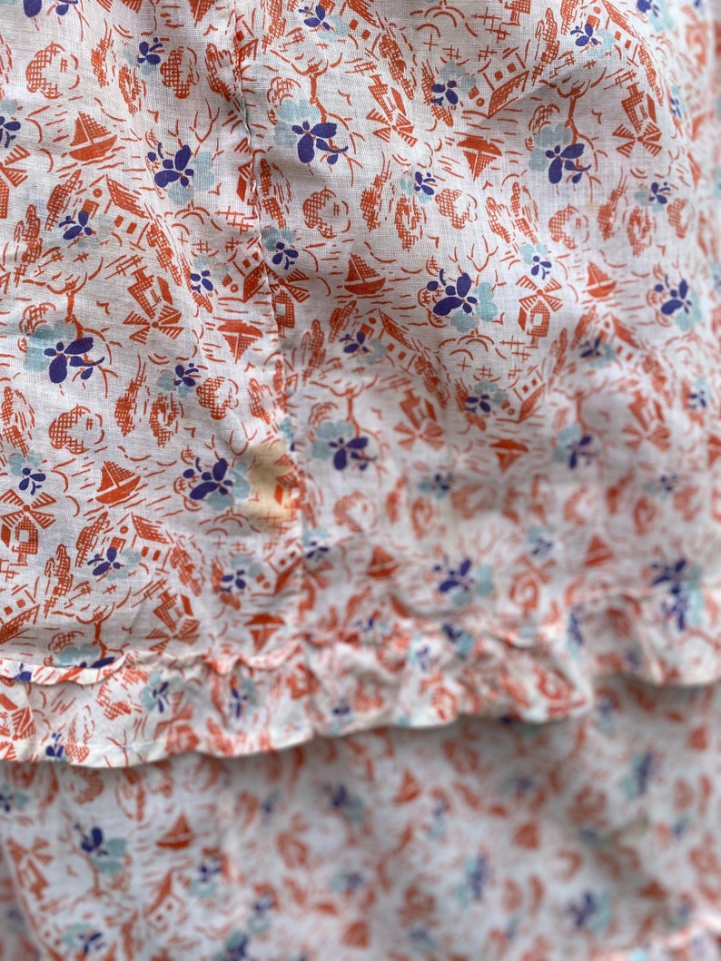 May include: White fabric with a repeating pattern of small orange houses, blue flowers, and sailboats. The fabric has a ruffled edge.
