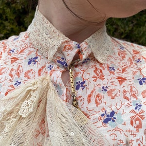 May include: A white blouse with a delicate lace collar and a gold chain necklace. The blouse has a floral print with red, blue, and white flowers. The lace is a delicate, intricate pattern.