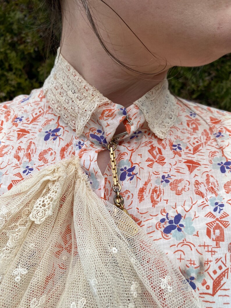 May include: A white blouse with a delicate lace collar and a gold chain necklace. The blouse has a floral print with red, blue, and white flowers. The lace is a delicate, intricate pattern.