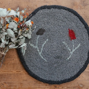 Op de afbeelding: Een rond grijs vloerkleed met een bloemenmotief. Het vloerkleed heeft een donkergrijze rand en bevat twee gestileerde bloemen met rode en groene accenten.