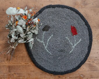 Puede incluir: Una alfombra redonda gris con un diseño floral. La alfombra tiene un borde gris oscuro y presenta dos flores estilizadas con acentos rojos y verdes.