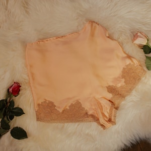 Puede incluir: Pantalones cortos de satén de color melocotón con un delicado ribete de encaje. Los pantalones cortos están sobre una superficie blanca y esponjosa.