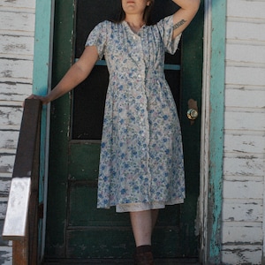 May include: A woman wearing a white dress with a floral pattern stands on a porch with a green door behind her. She is wearing brown boots and has her hand on her forehead.