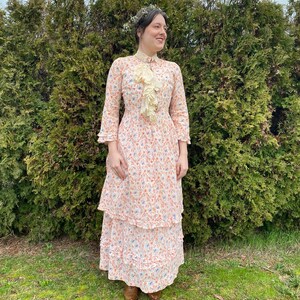 May include: A woman wearing a white dress with a floral print and a lace collar. The dress has a tiered skirt and is worn with brown shoes. She is standing in front of a green hedge.