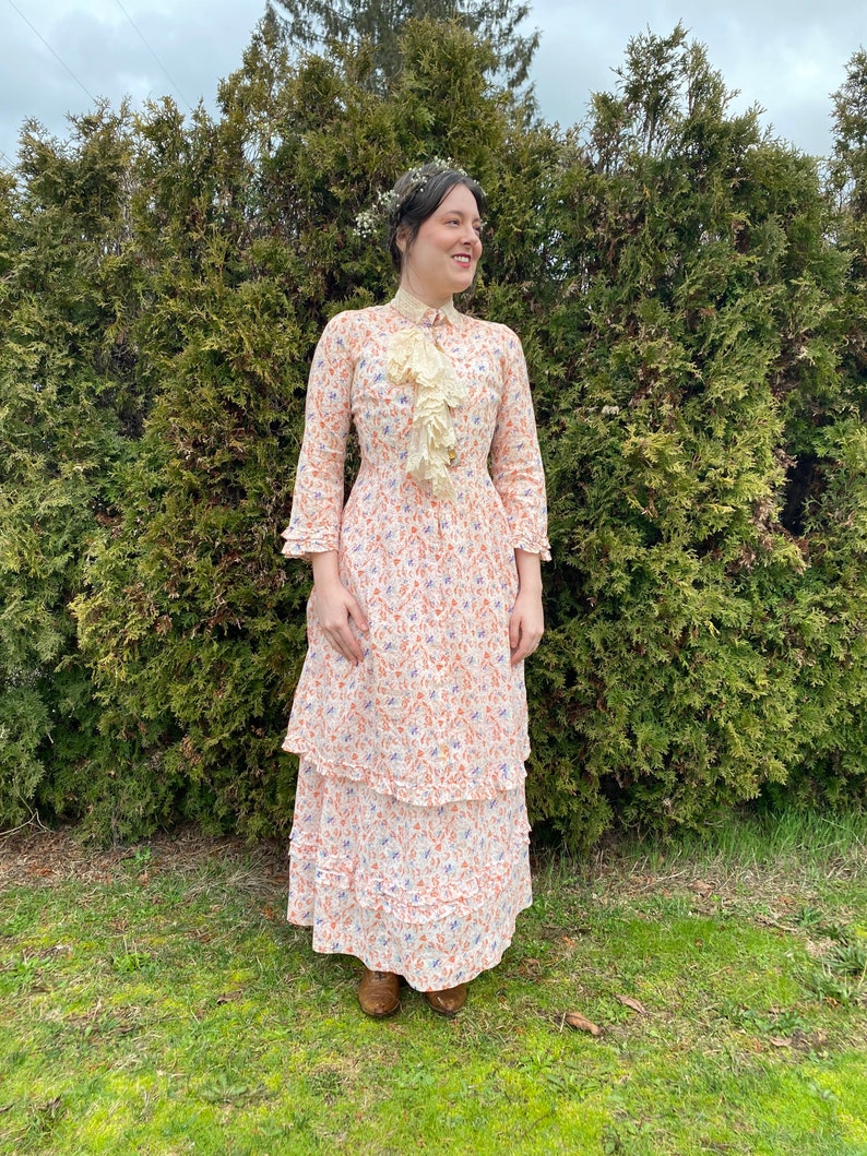 May include: A woman wearing a white dress with a floral print and a lace collar. The dress has a tiered skirt and is worn with brown shoes. She is standing in front of a green hedge.