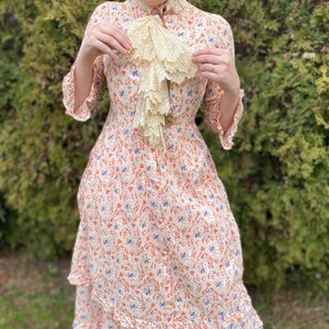 May include: A white dress with a floral print in shades of orange, blue, and white. The dress has a high neck and a ruffled collar. The dress is made of a lightweight fabric and has a tiered skirt.