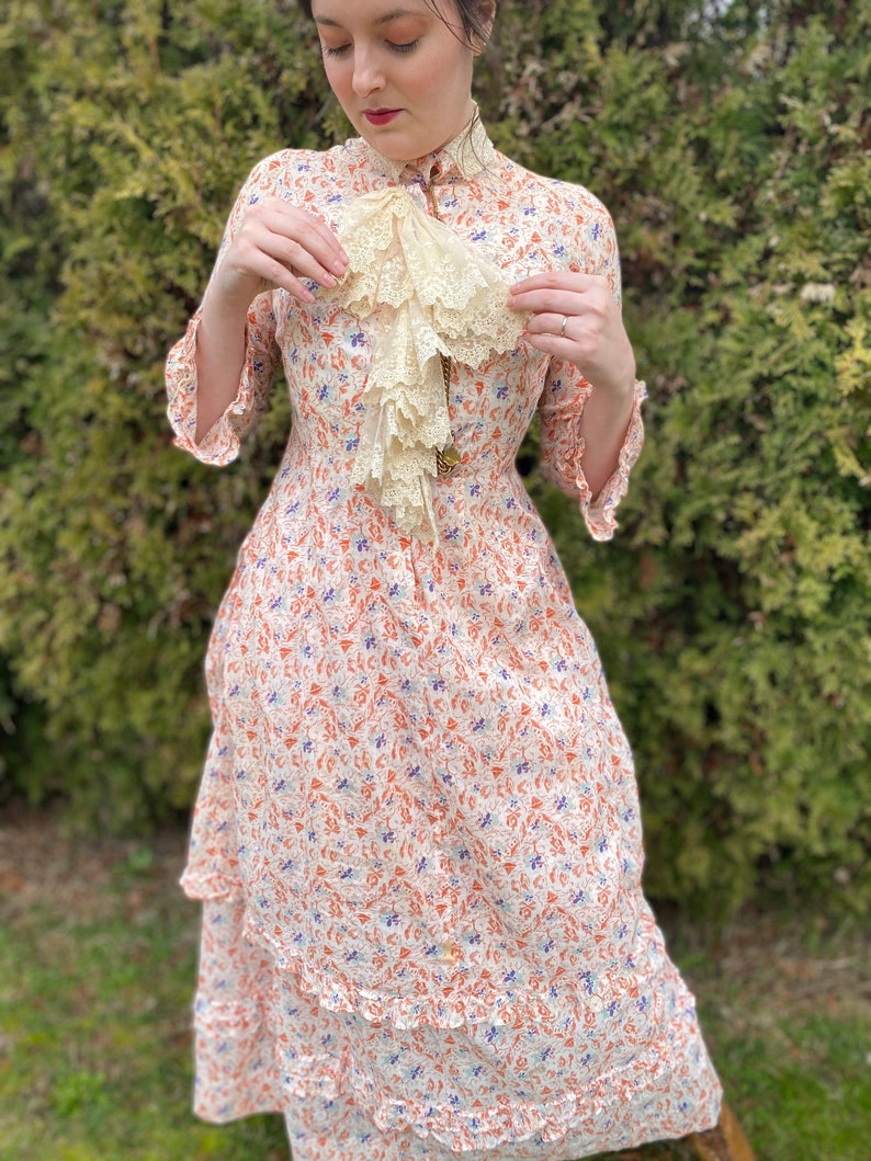 May include: A white dress with a floral print in shades of orange, blue, and white. The dress has a high neck and a ruffled collar. The dress is made of a lightweight fabric and has a tiered skirt.