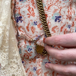 May include: A white dress with a floral print and a gold chain necklace with a decorative pendant. The dress has a lace trim on the left side.