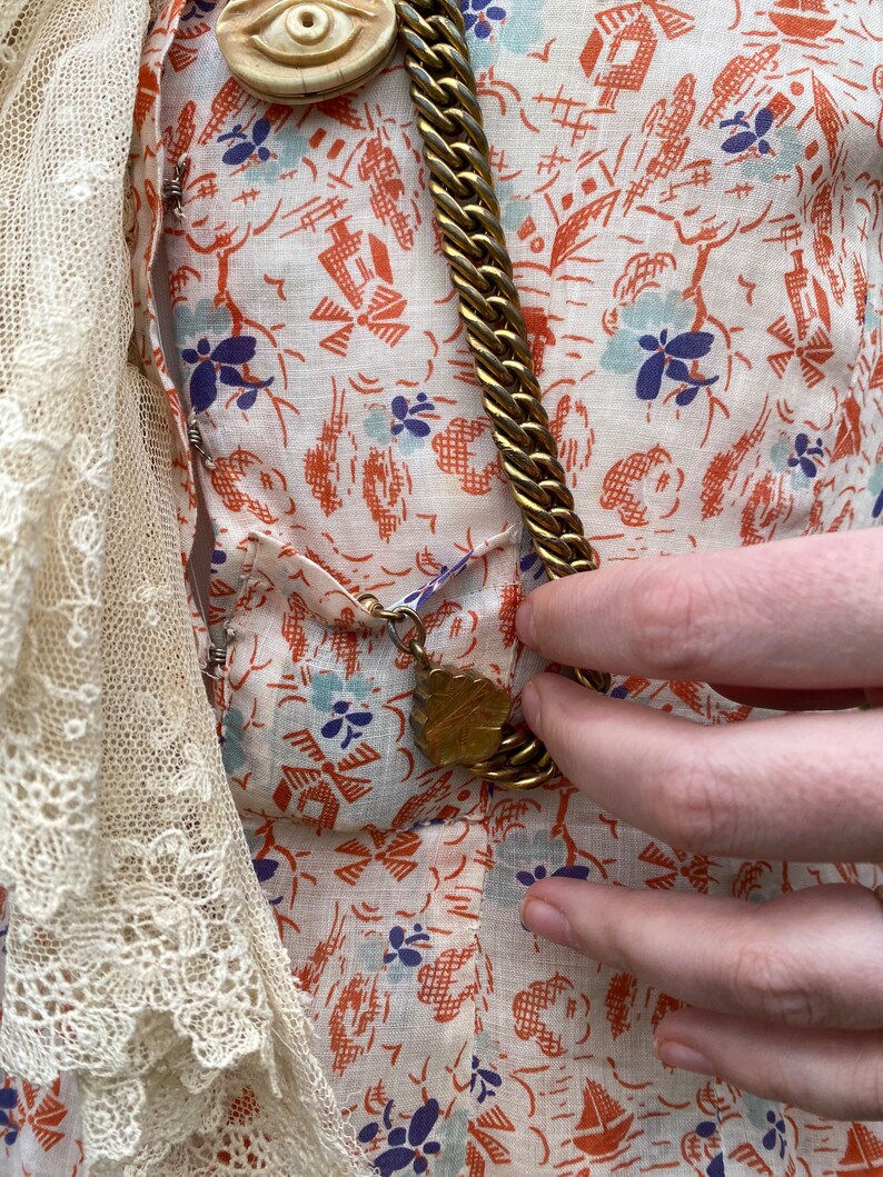 May include: A white dress with a floral print and a gold chain necklace with a decorative pendant. The dress has a lace trim on the left side.