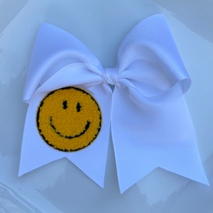 May include: White cheer bow with a yellow smiley face patch. The smiley face is fuzzy and has a black outline.