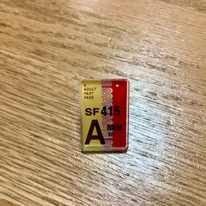 Op de afbeelding: Een gele en rode plastic pas met de tekst "A ADULT FAST PASS" en "SF415" erop gedrukt. De letter "A" is in grote zwarte letters gedrukt.