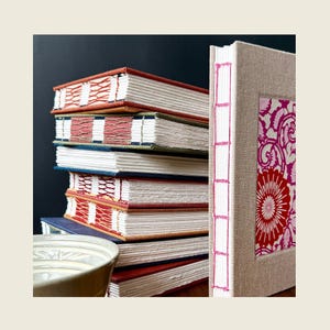 May include: A stack of six hardback books with exposed stitched spines in various colours, including orange, green, blue, and red. Beside the stack stands a single book with a beige fabric cover and a pink floral pattern on the front panel.