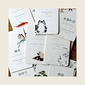 May include: A collection of rectangular white cards featuring Japanese kanji, English text, and small watercolor illustrations including a frog, an owl, a fish, and floral motifs. The cards display phrases like 'Rest in the space between', 'What needs to be said', and 'Some things are not yours to change'.