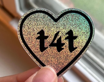 May include: A heart-shaped sticker with a holographic, glittery surface. The sticker has a black border and features the text "t4t" in a black, stylized font. The sticker is held by a person.