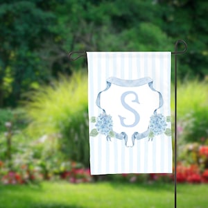 May include: A garden flag with a light blue and white striped background. The flag features a light blue letter "S" inside a decorative frame, with blue hydrangea flowers and ribbons. The flag is hanging on a black metal stand.