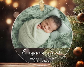 May include: A clear, round Christmas ornament with a photo of a newborn baby wrapped in a green blanket. The ornament includes the baby's name, birth date, time, and measurements: 20 inches, 8lbs 6oz. A gold ribbon is attached for hanging.