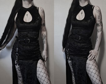 May include: A black velvet dress with a keyhole neckline and a high slit. The dress is accessorized with a black belt and chain details. The model is wearing fishnet stockings.