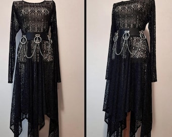 Op de afbeelding: Zwarte kanten jurk met lange mouwen en een asymmetrische zoom. De jurk is gestyled met een zwarte riem met zilveren ringen en kettingdetails. De jurk wordt op een mannequin getoond.