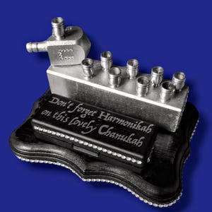 May include: Silver menorah with a spinning top on top. The menorah has eight candle holders and a base with the inscription "Don't forget Harmonikah on this lovely Chanukah".