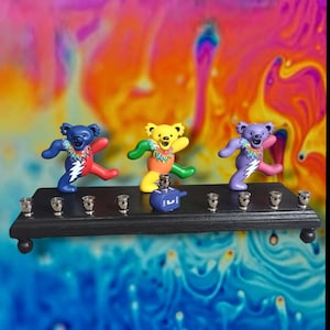 May include: A black wooden menorah with nine silver candle holders. Three colorful dancing bear figurines are placed on the menorah. The bears are blue, yellow, and purple with tie-dye shirts and lightning bolt designs. A blue dreidel is also on the menorah.