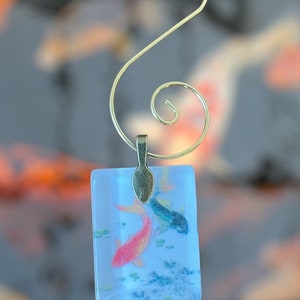 May include: A glass ornament featuring two koi fish swimming in a blue and white background. The ornament is attached to a gold-colored metal hook.