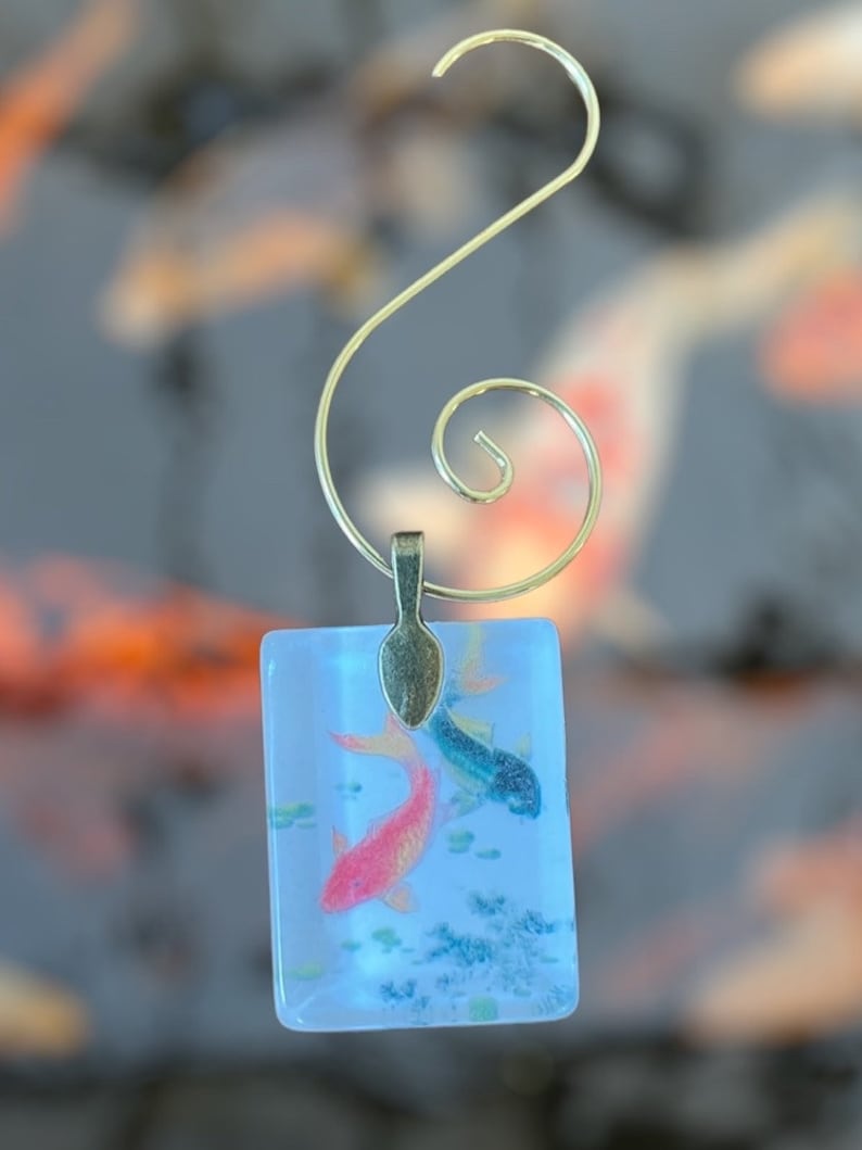 May include: A glass ornament featuring two koi fish swimming in a blue and white background. The ornament is attached to a gold-colored metal hook.