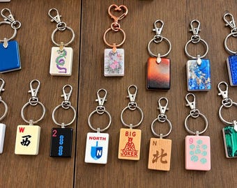 Op de afbeelding: Een verzameling van zeventien rechthoekige sleutelhangers met diverse ontwerpen, waaronder mahjongtegels, een dreidel met de tekst NORTH, een tegel met het opschrift BIG JOKER en tegels met bloem-, draak- en abstracte patronen.