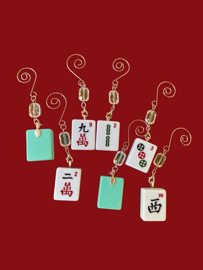 May include: Seven white and green mahjong tiles with Chinese characters and numbers, hanging from gold-colored wire loops with clear glass beads. The tiles are suspended against a red background.
