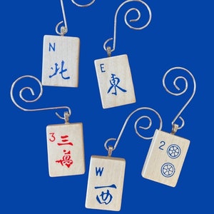 May include: Five wooden ornaments shaped like mahjong tiles with silver hangers. The tiles are painted with red, blue, and black characters and numbers. The tiles are labeled with the letters N, E, W, and the numbers 2 and 3.
