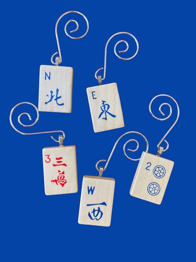 May include: Five wooden ornaments shaped like mahjong tiles with silver hangers. The tiles are painted with red, blue, and black characters and numbers. The tiles are labeled with the letters N, E, W, and the numbers 2 and 3.