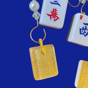 May include: Gold pendant earrings with a textured gold surface. The earrings are shaped like a square and have a small gold loop at the top. The earrings are hanging from a gold chain.