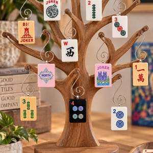 May include: A brown wooden tree-shaped display stand with a base labelled MAHJONG PLAY DECORATE GIFT. Ten mahjong tiles hang from silver metal hooks on the branches, featuring various designs including numbers, the word JOKER, and symbols.