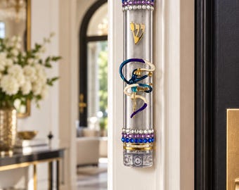 May include: A clear acrylic mezuzah case featuring a gold Hebrew letter Shin, a central blue and gold gymnast figure, and decorative bands of blue and purple rhinestones at the top and bottom, mounted vertically on a white door frame.