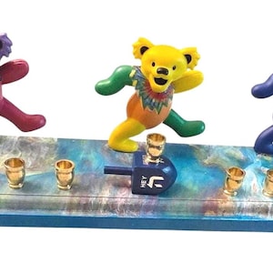May include: A vibrant Hanukkah menorah with three dancing bear figures in purple, yellow, and blue. The menorah has gold candle holders and a blue dreidel. The base is a mix of blue, white, and gold.