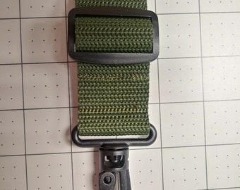 May include: A green nylon strap with a black plastic buckle and a black metal hook. The strap is designed for attaching to a bag or other object.