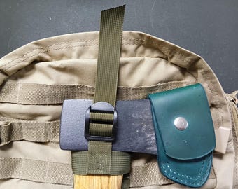 May include: A green leather sheath with a snap closure attached to a tan and green rucksack. A black and brown axe head is secured to the rucksack with a green strap and a black buckle.