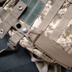 May include: Tan Gerber knife in a sheath attached to a digital camouflage backpack. The backpack has multiple straps and a Fieldline Tactical logo. The knife is secured with a strap and snap closures.
