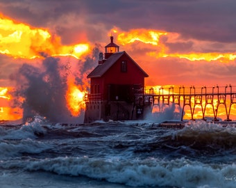 May include: A red lighthouse stands on a pier with crashing waves in the foreground. The sky is a fiery orange and yellow with clouds.