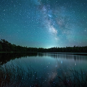 May include: A night sky filled with stars, including the Milky Way, reflected in a still lake. The trees surrounding the lake are silhouetted against the dark sky.
