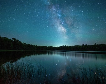 May include: A night sky filled with stars, including the Milky Way, reflected in a still lake. The trees surrounding the lake are silhouetted against the dark sky.