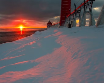 May include: A red lighthouse stands tall against a dramatic cloudy sky, with a bright orange sunset reflecting off the icy water. The lighthouse is connected to a pier with a wooden walkway, and the snow-covered ground is illuminated by the warm sunset glow.