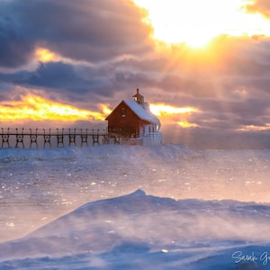 May include: A scenic photograph of a red lighthouse at the end of a pier, set against a dramatic sky. The sun shines brightly, casting golden rays through the clouds. The foreground features snow and ice, creating a winter landscape.