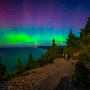 May include: A vibrant green and purple aurora borealis display over a dark blue lake with a wooden walkway leading up a grassy hill. The sky is filled with stars.