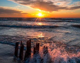 May include: A sunset over the ocean with a bright orange sky and clouds. The sun is setting behind the horizon, casting a golden glow on the water. There are some wooden posts in the foreground, partially submerged in the water. The waves are crashing against the posts, creating a spray of white foam.