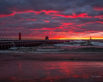 May include: A red lighthouse stands on a pier extending into a choppy sea. The sky is ablaze with a fiery red sunset, creating a dramatic scene.
