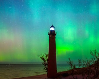 May include: A red brick lighthouse stands tall on a sandy beach under a vibrant green and blue aurora borealis. The lighthouse has a white light shining from the top. The sky is filled with stars.